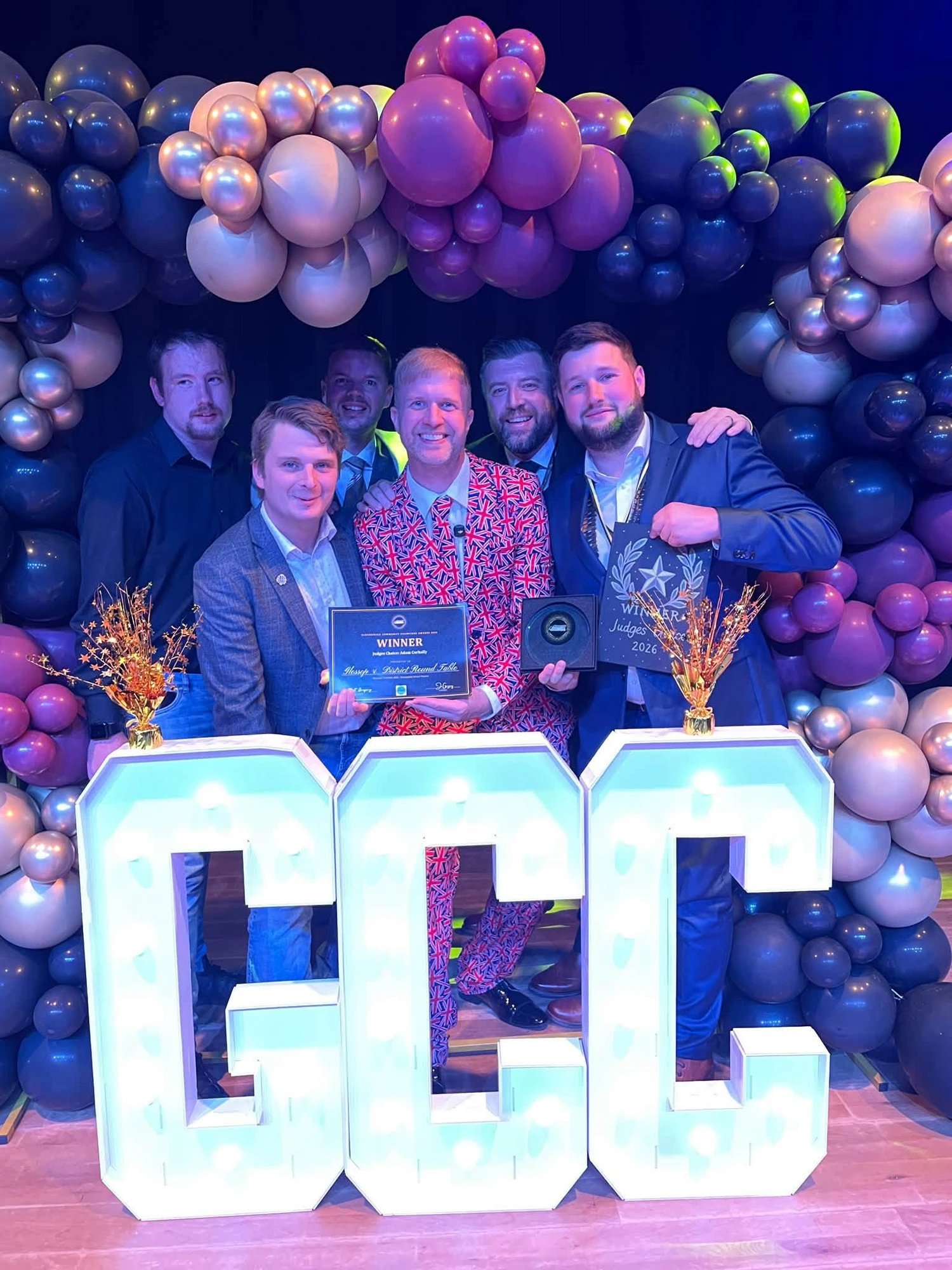 Glossop & District Round Table members on stage at the Glossopdale Community Champions Awards 2026, holding the WINNER certificate for the Adam Corbally Judges' Choice Award in front of giant light-up GCC letters
